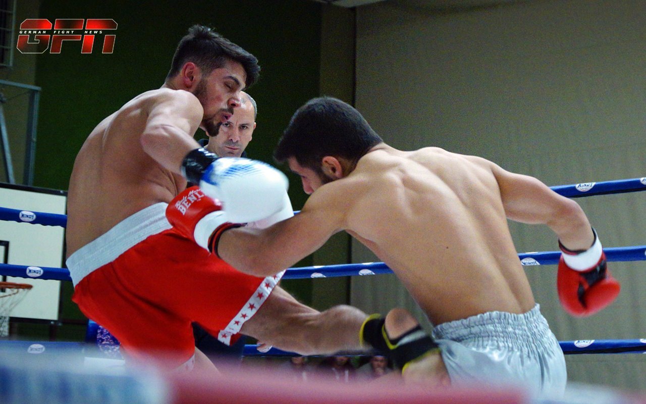 Zwei K&auml;mpfer im Ring, einer f&uuml;hrt einen Kick aus, w&auml;hrend der andere defensiv ist. Ein Schiedsrichter beobachtet das Geschehen.