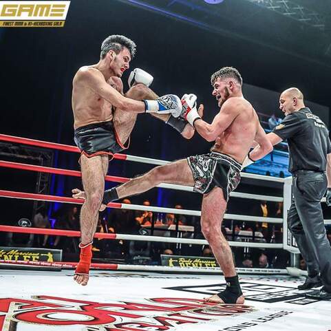 Zwei K&auml;mpfer im Ring bei einem Kickboxkampf, einer hebt das Knie zur Abwehr, w&auml;hrend der Schiedsrichter zuschaut.