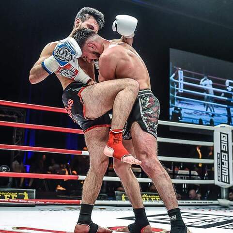 Zwei K&auml;mpfer im Clinch w&auml;hrend eines Wettkampfs im Ring, einer hebt den anderen an, w&auml;hrend Zuschauer im Hintergrund blitzen.