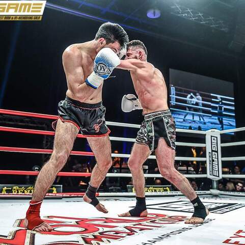 Zwei K&auml;mpfer im Ring, einer schl&auml;gt mit einem Jab, w&auml;hrend der andere in Deckung geht. Hintergrundbeleuchtung und Zuschauer sichtbar.