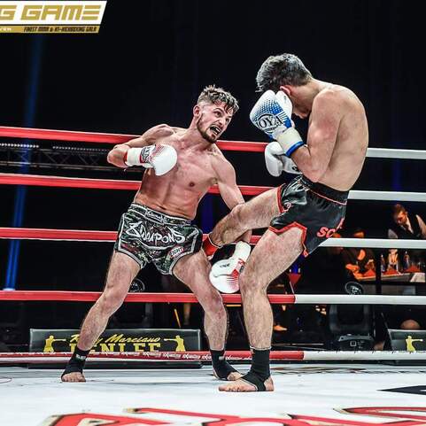 Zwei K&auml;mpfer in Action im Boxring, einer tritt zu seinem Gegner, w&auml;hrend der andere mit Handschuhen kontert.