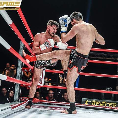 Zwei K&auml;mpfer im Ring, einer f&uuml;hrt einen Kick gegen den Gegner, umgeben von roten Seilen und Zuschauern im Hintergrund.