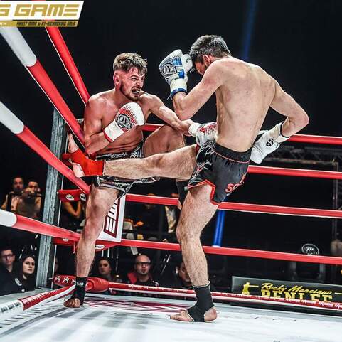 Zwei K&auml;mpfer im Ring, einer f&uuml;hrt einen Kick aus, w&auml;hrend der andere defensiv reagiert. Rote Ringseile umgeben die Szene.