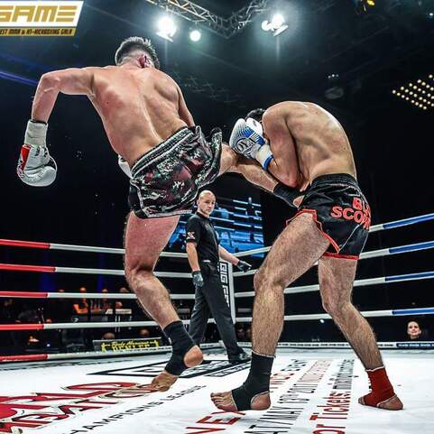 Zwei K&auml;mpfer im Ring, einer f&uuml;hrt einen Kick aus, w&auml;hrend ein Schiedsrichter und Zuschauer im Hintergrund zu sehen sind.