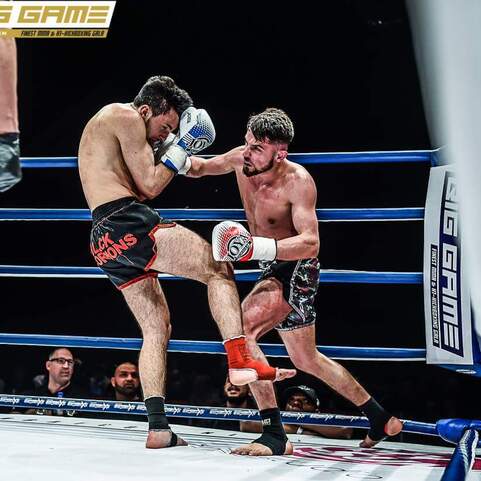 Zwei K&auml;mpfer stehen in einem Ring, einer schl&auml;gt einen K&ouml;rpersto&szlig;, Zuschauer sind im Hintergrund sichtbar.