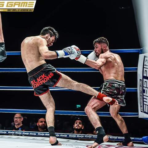 Zwei K&auml;mpfer im Boxring, einer tritt aus einer Offensive, w&auml;hrend der andere sich abwehrt. Zuschauer sind im Hintergrund sichtbar.