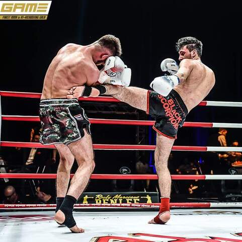 Zwei K&auml;mpfer im Ring, einer f&uuml;hrt einen Kick aus, w&auml;hrend der andere sich verteidigt. Rote Seile umrahmen die Kampffl&auml;che.