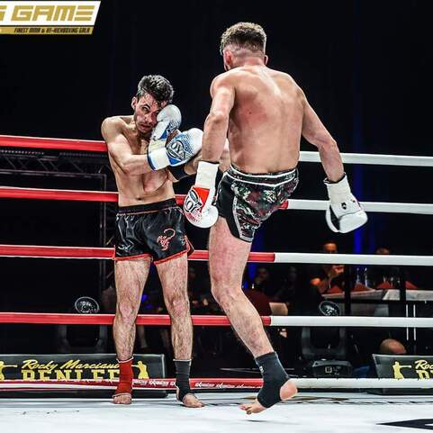 Zwei K&auml;mpfer im Ring, einer blockt einen Schlag, der andere hebt das Bein zum Kick. Rote Seile umgeben den Wettkampfort.