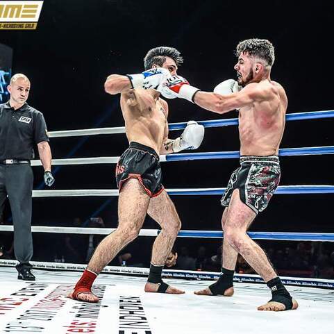 Zwei K&auml;mpfer im Ring w&auml;hrend eines Matches, einer f&uuml;hrt einen Schlag, ein Schiedsrichter beobachtet das Geschehen.