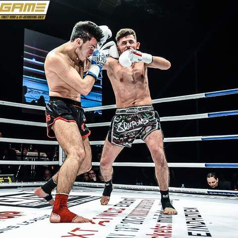 Zwei Kickboxer im Ring, einer trifft den anderen mit einem Schlag, Hintergrund unscharf, Zuschauer sichtbar.