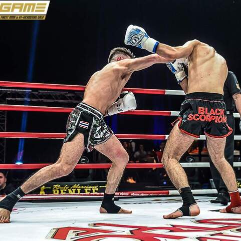 Zwei K&auml;mpfer im Ring, einer setzt einen linken Punch gegen den anderen ein, w&auml;hrend Zuschauer im Hintergrund beobachtet.