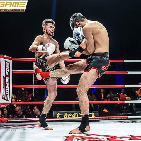 Zwei K&auml;mpfer im Ring, einer hebt das Bein zum Kick, w&auml;hrend der andere sich verteidigt. Rote Seile um den Ring.