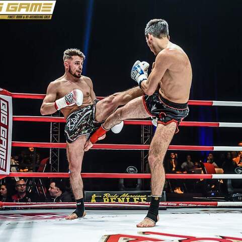 Zwei K&auml;mpfer im Ring f&uuml;hren einen Kick im Muay Thai Wettkampf aus, Zuschauer sind im Hintergrund sichtbar.