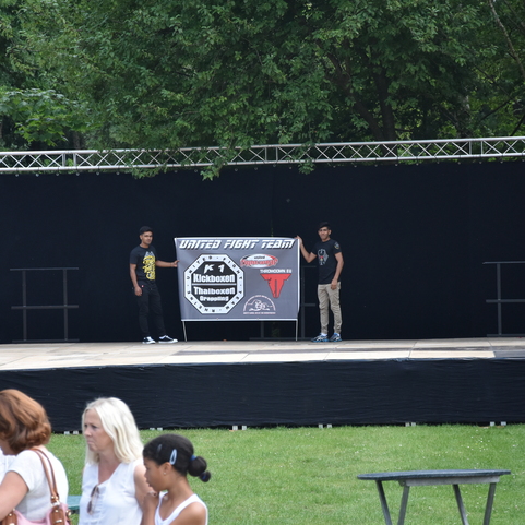 Zwei junge M&auml;nner stehen auf einer leeren B&uuml;hne vor einem Banner, umgeben von Menschen im Freien.