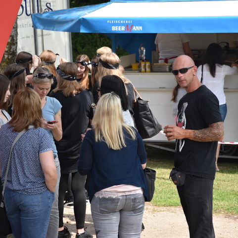 Menschenmenge vor einem Imbissstand, einige in Wartestellung, andere mobil mit Handys, im Freien bei sonnigem Wetter.