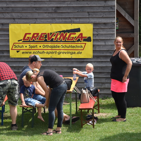 Gruppenszene mit Familien am Tisch, ein Banner mit &bdquo;Grevinga&ldquo; im Hintergrund, im Freien auf gr&uuml;ner Wiese.