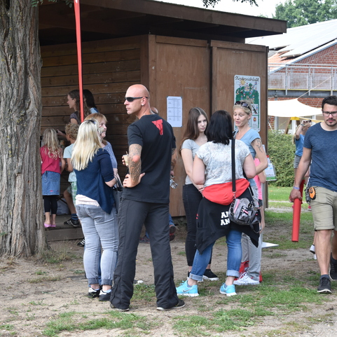 Menschenmenge vor einem Holzgeb&auml;ude, einige unterhalten sich, w&auml;hrend andere an einem Tisch stehen und Informationen einholen.