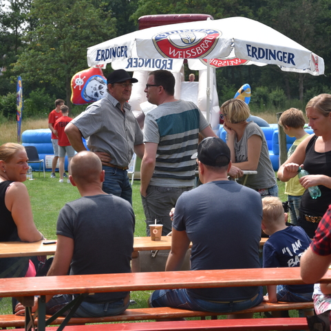 Personen stehen an einem Tisch mit Getr&auml;nken unter Schirmen, im Hintergrund ein Spielebereich auf einem Festplatz.