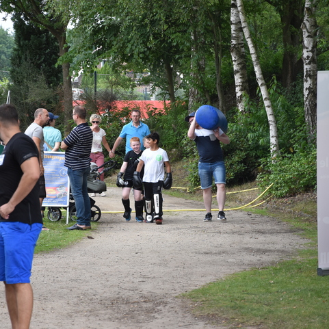 Menschen flanieren auf einem Weg, w&auml;hrend ein Junge einen blauen Ball tr&auml;gt und andere Kinder in Schutzausr&uuml;stung laufen.