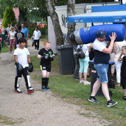 Gruppe von Kindern in Boxkleidung mit einem Boxsack auf einem Sportgel&auml;nde, umgeben von Zuschauern und St&auml;nden.