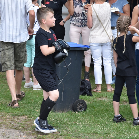 Junge in schwarzer Kleidung mit Boxhandschuhen steht neben einer Gruppe von Menschen auf einer Wiese.