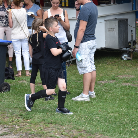 Ein Junge in Sportkleidung tr&auml;gt Handschuhe und l&auml;uft &uuml;ber eine Wiese, im Hintergrund stehen Menschen und M&uuml;lltonnen.
