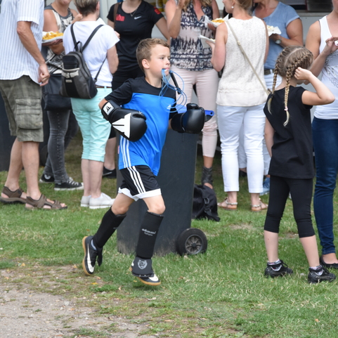 Ein Junge in Boxhandschuhen und Trikot l&auml;uft auf einer Wiese, umgeben von Zuschauern in Freizeitkleidung.