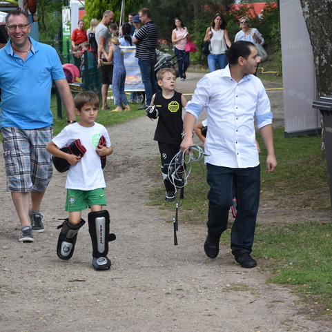 Familien und Kinder gehen auf einem Waldweg, ein Junge tr&auml;gt Schutzausr&uuml;stung und h&auml;lt einen Schl&auml;ger in der Hand.