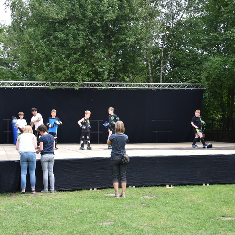 Gruppe von Kindern, die auf einer B&uuml;hne verschiedenen Sportarten demonstrieren, w&auml;hrend Zuschauer zuschauen.