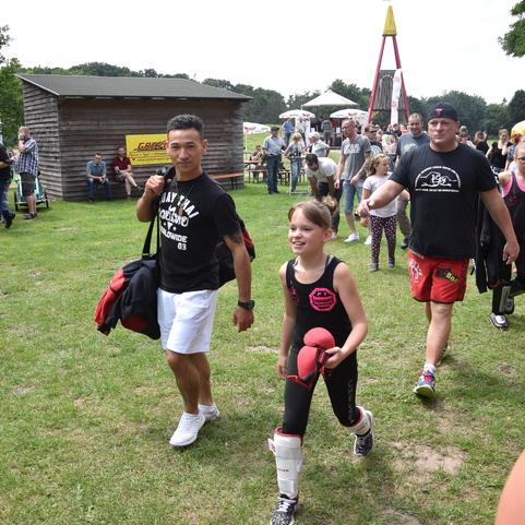 Drei Personen gehen auf einer Veranstaltung mit Sporttaschen, w&auml;hrend Zuschauer auf B&auml;nken sitzen. Gr&uuml;nen Platz im Hintergrund.