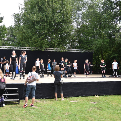 Gruppen von Menschen nehmen an einer Fitnessveranstaltung auf einer B&uuml;hne im Freien teil, umgeben von B&auml;umen.