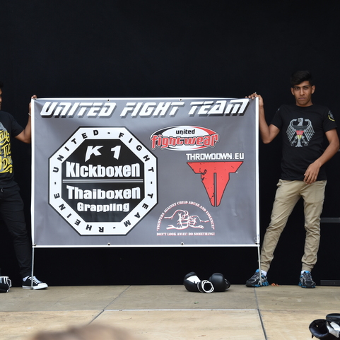 Zwei Jugendliche halten ein Banner der "United Fight Team" mit Informationen zu Kampfsportarten auf einer B&uuml;hne.