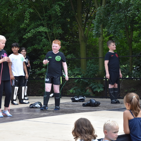 Gruppe von Kindern in Sportbekleidung beim Training auf einer Freiluftfl&auml;che, einige Zuschauer sitzen am Rand.