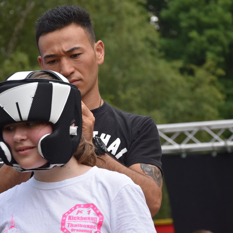 Trainer stattet eine Sch&uuml;lerin mit einem Kopfschutz f&uuml;r das Kickboxen aus, w&auml;hrend im Hintergrund eine Veranstaltung stattfindet.