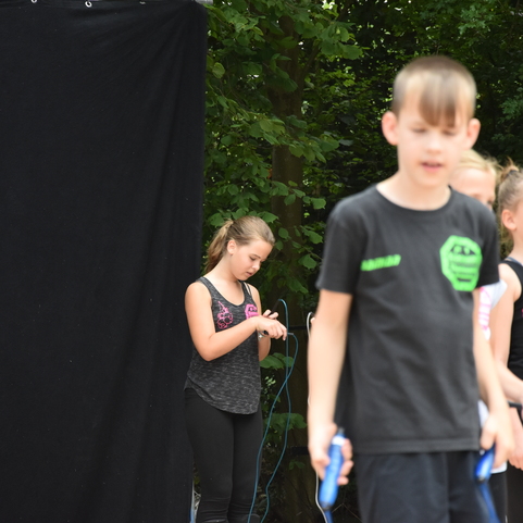 M&auml;dchen in Sportbekleidung springt Seil auf einer B&uuml;hne mit schwarzem Hintergrund, andere Kinder beobachten.