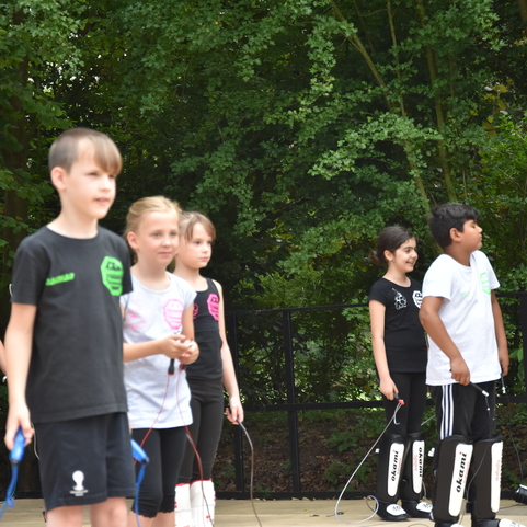Sechs Kinder in Sportkleidung stehen auf einer B&uuml;hne, einige mit Seilen, umgeben von B&auml;umen.