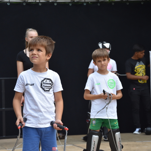Zwei Kinder mit Springseilen stehen auf einem Trainingsgel&auml;nde, im Hintergrund ein Boxsack und eine Bannermitteilung.