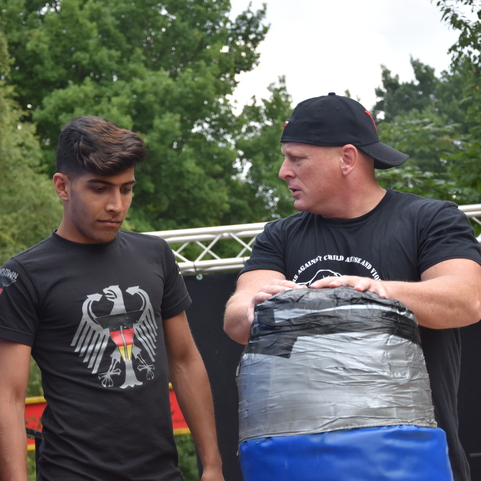 Zwei M&auml;nner stehen auf einem Sportplatz, einer h&auml;lt einen gepolsterten Schlagpolster, der andere schaut konzentriert.