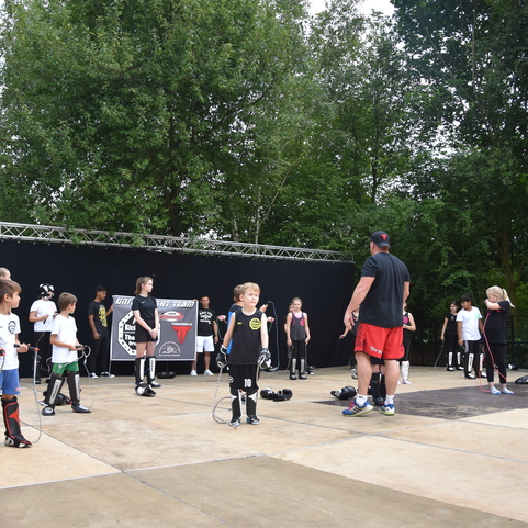 Gruppe von Kindern mit Schutzausr&uuml;stung beim Training auf einer B&uuml;hne im Freien, angeleitet von einem Trainer.