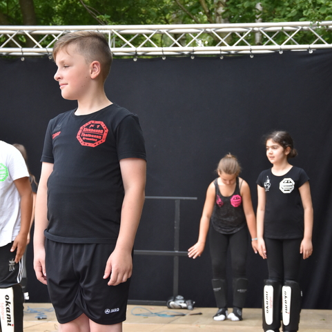 Gruppe von Kindern in Sportbekleidung auf einer B&uuml;hne, bereit f&uuml;r eine Auff&uuml;hrung vor schwarzem Hintergrund.