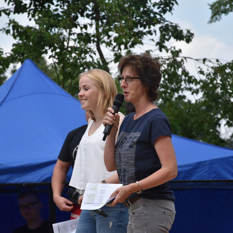 Zwei Frauen stehen vor einem blauen Zelt, eine spricht mit Mikrofon, die andere l&auml;chelt und h&ouml;rt zu.