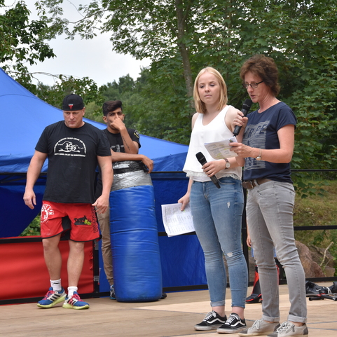Zwei Frauen mit Mikrofonen stehen auf einer B&uuml;hne, im Hintergrund sieht man einen Boxsack und Zuschauer.