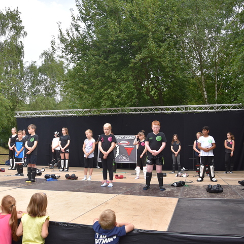 Gruppe von Kindern in Sportkleidung steht auf einer B&uuml;hne, Zuschauer sitzen davor, umgeben von B&auml;umen.