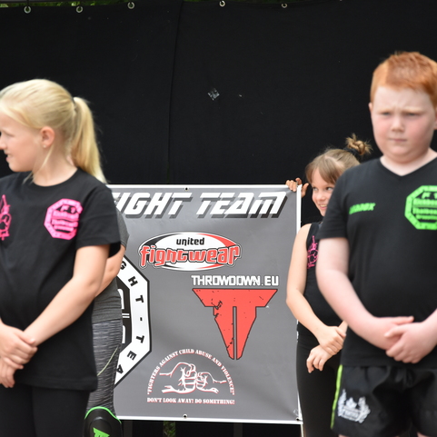Drei Kinder in Schwarz stehen vor einem Banner der &bdquo;United Fightwear&ldquo; auf einer Sportveranstaltung.