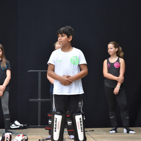 Gruppe von Kindern in Sportkleidung steht auf einer B&uuml;hne, einige tragen Sch&uuml;tzer, vor schwarzem Hintergrund.