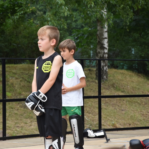 Zwei Jungen in Sportkleidung stehen auf einer Trainingsfl&auml;che, w&auml;hrend eine junge Frau im Hintergrund zuschaut.