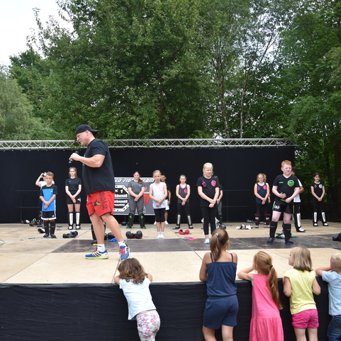 Gruppe von Kindern in Sportkleidung &uuml;bt auf einer B&uuml;hne, w&auml;hrend ein Trainer Anweisungen gibt. Zuschauer beobachten von vorne.