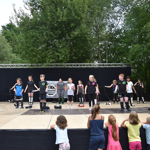 Kinder in Sportkleidung f&uuml;hren eine Gruppen&uuml;bung auf einer B&uuml;hne vor, w&auml;hrend einige Zuschauer im Vordergrund zuschauen.