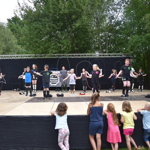 Kinder springen Seil auf einer B&uuml;hne, w&auml;hrend eine Gruppe von Zuschauern im Vordergrund zuschaut. B&auml;ume im Hintergrund.