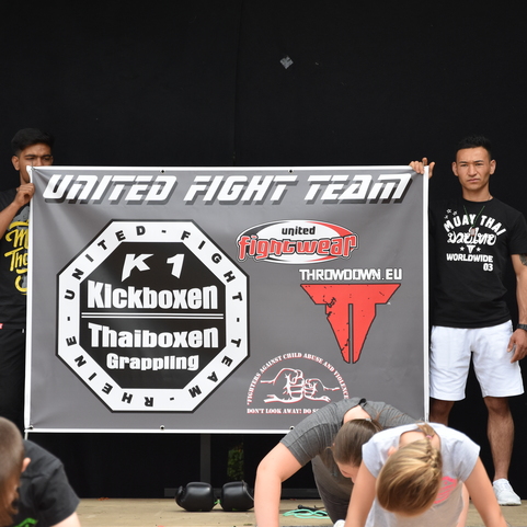 Zwei Personen halten ein Banner des United Fight Teams, w&auml;hrend im Vordergrund Teilnehmer in Sportkleidung trainieren.